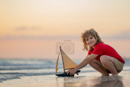 Téléchargez les photos : Petit garçon jouant avec un voilier jouet, un bateau jouet. Concept voyage et aventure. Enfant se sentant aventureux en croisière. Enfant sur la plage de coucher de soleil d'été. Enfant jouant sur la plage au coucher du soleil - en image libre de droit