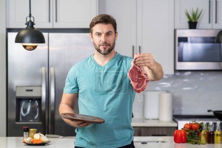 Téléchargez les photos : Charmant homme cuisinant du steak de bœuf dans la cuisine. Portrait d'un homme occasionnel cuisinant avec des ingrédients de viande. Homme décontracté préparant de la viande crue dans la cuisine. Chef cuisinier steak de boeuf - en image libre de droit