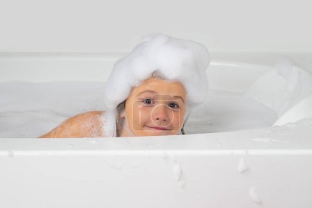 Foto de Niños bañándose. Niño lavándose con una burbuja en el baño. Lindo niño se baña, acostado en un baño blanco con espuma de jabón y champú - Imagen libre de derechos