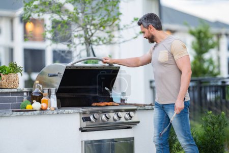 Téléchargez les photos : Beau 40s homme préparant barbecue. Cuire la viande masculine sur le barbecue grill. Guy cuisinant de la viande sur le barbecue pour le dîner familial d'été dans la cour arrière de la maison - en image libre de droit