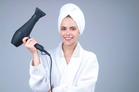 Téléchargez les photos : Femme en peignoir et serviette sur la tête avec sèche-cheveux en studio. Portrait de modèle féminin avec sèche-cheveux. Soins capillaires et beauté. Routine matinale - en image libre de droit
