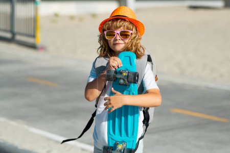 Téléchargez les photos : Petit garçon 7-8 ans tenir skateboard posant sur la rue en plein air. Concept de style de vie de l'enfance. Mode enfant en chemise d'été chapeau élégant et lunettes de soleil - en image libre de droit