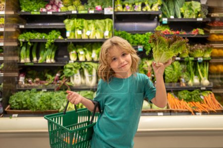 Téléchargez les photos : Enfant avec salade de laitue. Le gamin choisit des légumes et des fruits frais dans le magasin. Enfant achetant de la nourriture dans un supermarché - en image libre de droit