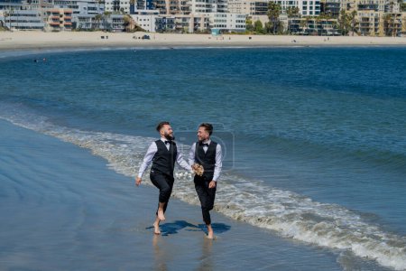 Téléchargez les photos : Grooms gays marchant ensemble sur la plage de la mer pendant le jour du mariage. Hommes romantiques dans l'eau de mer. Portrait de couple gay amoureux le jour du mariage. mariage gay en plein air - en image libre de droit