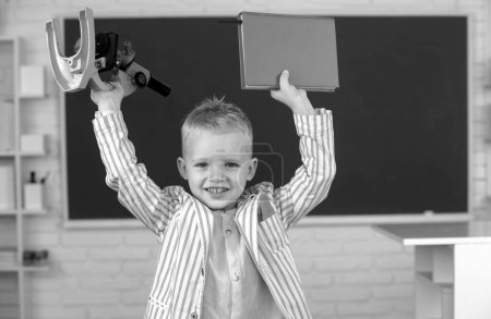 Photo for Amazed school boy with microscope and book. Funny face of little student of primary school study in classroom at school - Royalty Free Image