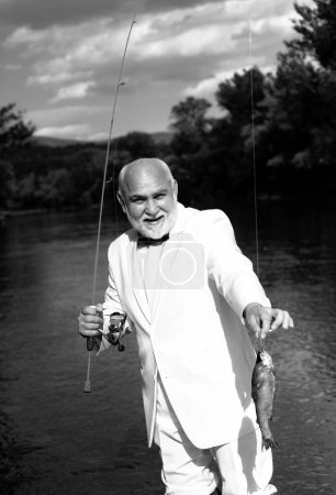 Téléchargez les photos : Un pêcheur a attrapé un poisson de truite. Portrait d'homme âgé joyeux pêche. Grand-père avec des poissons de pêche. Homme mûr pêcheur en costume blanc et noeud papillon avec canne à pêche, bobine filante sur la rivière - en image libre de droit