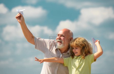 Téléchargez les photos : Enfant garçon et grand-père avec avion en papier sur ciel bleu et nuages. Hommes génération grand-père et petit-enfant. Aîné parent âgé avec enfant - en image libre de droit