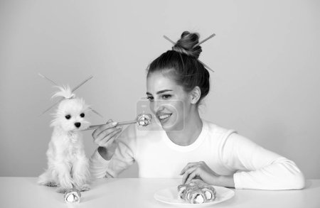 Téléchargez les photos : Fun jeune femme avec chiot animal manger des sushis sur fond gris. Gros plan portrait de beauté femme et chien manger des rouleaux de sushi - en image libre de droit