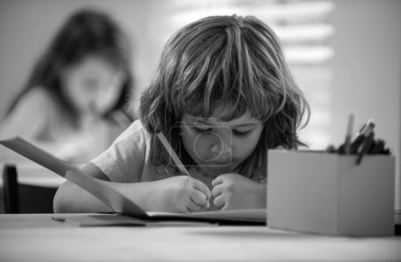 Photo for School kid writing something in copybook and sitting at table in classroom. Back to school. Happy smiling pupil drawing at the desk. Child from primary school in the classroom - Royalty Free Image