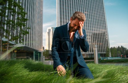 Photo for Stressed businessman sitting on grass outside office after project failure. Confused young man frustrated by business problem - Royalty Free Image