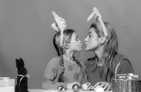 Atractiva mujer joven con niña linda se están preparando para la Pascua. Madre e hija con orejas de conejo se besan pasando tiempo juntas antes de Pascua mientras pintan huevos
