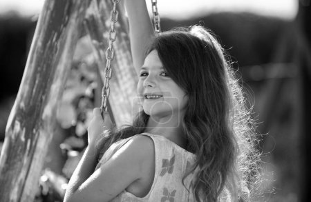Téléchargez les photos : Happy child girl laughing and swinging on a swing at the park in summer. Kids happiness and smile. Child playing on outdoor playground. Close up portrait of a beautiful child girl dreaming - en image libre de droit