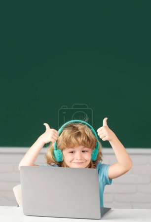 Foto de Lindo chico con pulgares arriba usando auriculares, estudio con computadora portátil en el aula, escuchando curso de lección de audio. Primer día en la escuela - Imagen libre de derechos