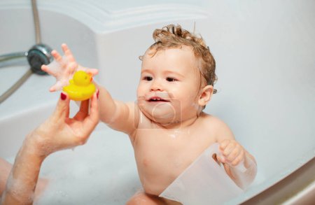 Téléchargez les photos : Un gamin se baignant dans une baignoire. Bains de bébé heureux drôles dans la baignoire avec de l'eau et de la mousse. Hygiène des enfants. Enfant souriant dans la salle de bain avec canard jouet - en image libre de droit