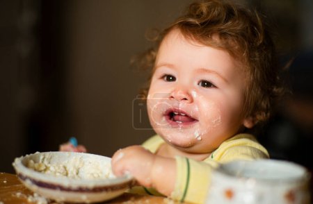 Photo for Cheerful smiling child child eats itself with a spoon Baby eating with dirty face. Smiling little child eating food on kitchen. Kid hungry so parent feed him - Royalty Free Image
