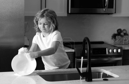 Téléchargez les photos : Enfant femme de ménage laver la vaisselle sur de l'eau savonneuse. Mignon garçon drôle lavant la vaisselle dans la cuisine. Un gamin américain qui apprend les tâches domestiques à la maison. Nettoyage vaisselle pendant les travaux ménagers - en image libre de droit