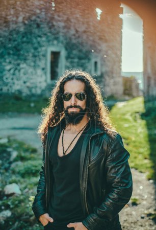 Stylish young fashionable man with long wavy hair wearing bikers leather jacket and sunglasses. Self confident handsome man in sunglasses. Macho man walking outside alone