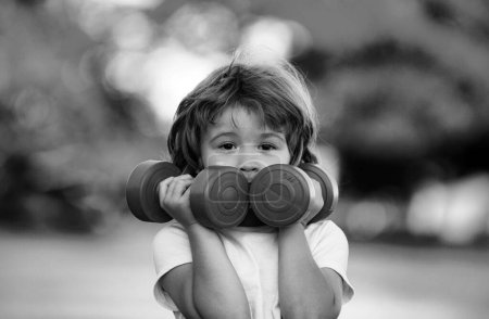 Photo for Cute funny little boy doing exercises with dumbbells in green park. Closeup portrait of sporty child with dumbbells. Happy child boy exercising outdoor. Healthy activities kids lifestyle - Royalty Free Image