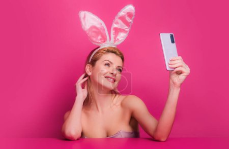 Téléchargez les photos : Sexy femme lapin de Pâques avec des oreilles de lapin ayant appel tenant téléphone intelligent dans la main tir selfie sur la caméra avant. isolé sur rose - en image libre de droit