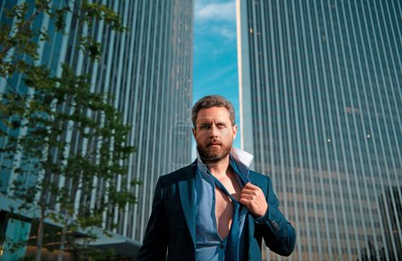 Photo for Sexy businessman. Confident young man in suit standing outdoors with cityscape in the city background. Close up portrait of exhausted frustrated stressed sad upset entrepreneur trying to take off tie - Royalty Free Image