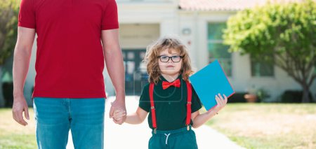 Photo for Parent and pupil of primary school go hand in hand. Teacher in t-shirt and cute schoolboy with backpack near school park - Royalty Free Image