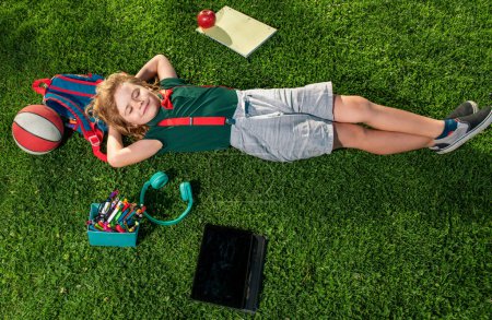 Foto de Niño feliz disfrutando en el parque durante el día de verano y otoño en la naturaleza en la hierba verde con útiles escolares al aire libre. Vista superior del niño tendido sobre hierba verde - Imagen libre de derechos