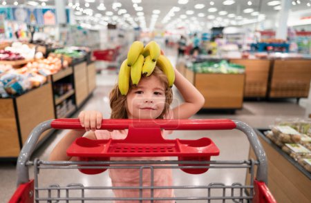 Téléchargez les photos : Enfant drôle avec panier achetant de la nourriture à l'épicerie ou au supermarché. Des gamins surpris. Banane sur drôle tête d'enfant - en image libre de droit