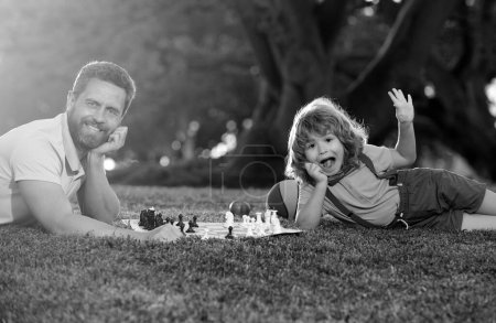 Téléchargez les photos : Père et fils jouant aux échecs allongés sur l'herbe à Lawn Park. Jeux et animations pour enfants. Concept de famille. Fête des Pères, famille amoureuse, parentalité, concept d'enfance - en image libre de droit