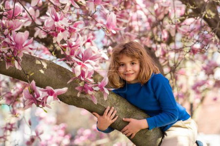 Kleines Kind riecht Frühlingsblume im Freien. Porträt eines lächelnden Kindergesichts in der Nähe blühender Frühlingsblumen. Kind zwischen Zweigen des Frühlingsbaums in Blüten. Nettes Kindergesicht umgeben von Frühlingsblüte