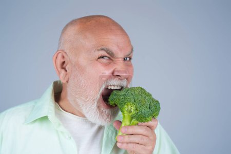 Foto de Divertido Senior en Dieta. Brócoli. Viejo hombre maduro comiendo brócoli. Comida saludable, dieta para personas mayores. Hombre maduro muerde brócoli en el estudio. Hombre vegano. Dieta Envejecida. Comida saludable. Brócoli para bajar de peso - Imagen libre de derechos