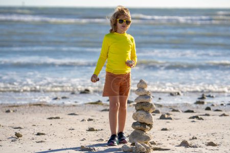 Téléchargez les photos : Petit garçon jouant avec des pierres sur la plage. Les enfants jouent avec la pyramide de pierres sur la plage, le paysage marin, le repos et les vacances d'été des enfants de bord de mer. Enfant jouant sur la plage et construisant tour en pierre - en image libre de droit