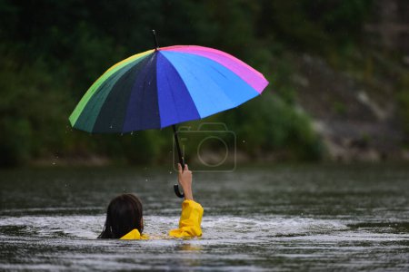 Foto de Concepto de inundación. Día de lluvia. Una mujer divertida con impermeable sostiene el paraguas en el agua. Chica bajo la lluvia con paraguas en temporada de lluvias, Deluge. Inundación. Clima lluvioso e inundaciones. Tormenta con ella - Imagen libre de derechos