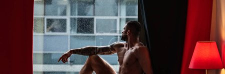 Foto de Hombre ventana en tiempo lluvioso mirando a través de la ventana con gotas de lluvia, bandera de otoño. Hombre musculoso guapo en el dormitorio en cortinas de ventana. Joven guapo sexy hombre descansando en casa - Imagen libre de derechos