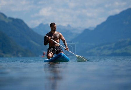 Sommerurlaub in den Schweizer Alpen. Mann paddelt auf Tretbrett oder im See. Sommerlicher Lebensstil. Männliche Fit-Spot-Modell Schwimmen mit Paddelbrett