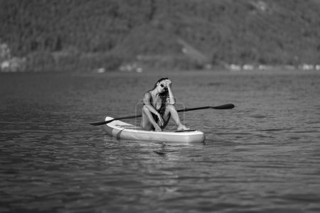 Fit woman with paddle board. Woman paddling on paddleboard. Sexy woman on sup board paddle surfing. SUP surfing in summer vacation in Alps lake in Switzerland. Summer black and white photo