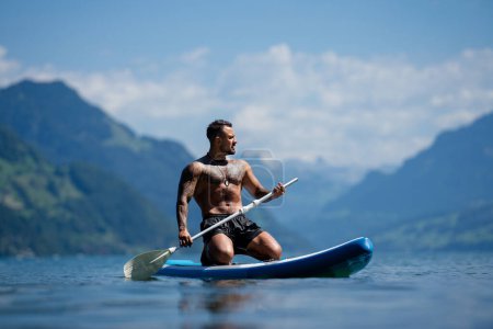 Téléchargez les photos : Vacances d'été dans les Alpes suisses. Homme pagayant sur la planche à pagaie ou soup dans le lac des Alpes. Style de vie estival. Modèle masculin spot avec paddle board - en image libre de droit