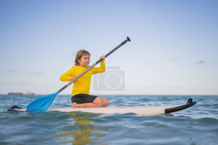 Foto de Un chico montando en una tabla de remo. Vacaciones de verano. Niño surfista de paddle surf niño rubio en la playa de verano. Niño feliz flotando en un paddleboard en el mar - Imagen libre de derechos