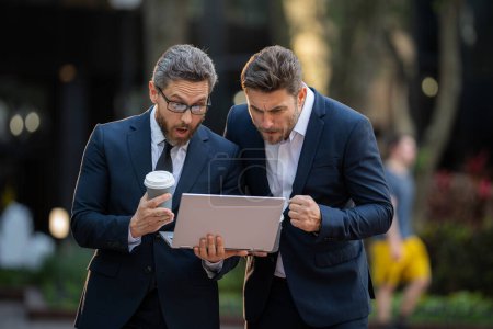 Two handsome young businessmen in classic suits using laptop. Business men team using laptop outdoor. Businessmen looking laptop with their business success in city. Two business teams success