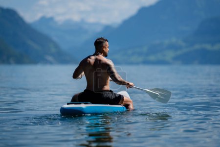 Téléchargez les photos : Homme sexy pagayant sur le paddleboard. Mode de vie sain en été. Sport d'été. SUP surf tour en aventure vacances d'été dans les Alpes lac en Suisse. Mâle ajustement musculaire modèle natation avec planche à pagaie - en image libre de droit