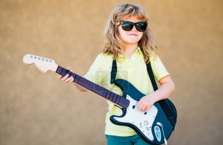 Téléchargez les photos : Enfant drôle avec des cheveux bouclés blonds jouant de la guitare sur fond jaune beige. Garçon guitariste - en image libre de droit