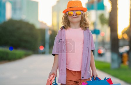 Photo for Fashion kid with shopping bags. Child in trendy hat and shirt shopping near shopping center. Happy boy holding shopping bags at the mall. Funny little customer. Street outfit, kids sales - Royalty Free Image