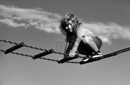 Photo for Cute boy climbs up the ladder on the playground. Child climbs up the ladder against the blue sky. Beautiful smiling cute boy on a playground. Kids activities - Royalty Free Image