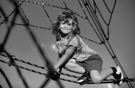 Téléchargez les photos : Joli gosse souriant escaladant le filet à l'aire de jeux. Parc à cordes pour enfants. Visage drôle d'enfants - en image libre de droit