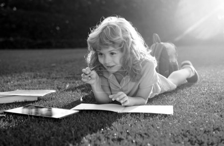 Téléchargez les photos : Un gamin lisant un livre allongé sur l'herbe. Mignon petit enfant en vêtements décontractés avec un crayon écrit sur le carnet et souriant tout en étant allongé sur l'herbe dans le parc. Nerd petit génie, éducation et apprentissage intelligent - en image libre de droit