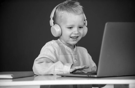 Téléchargez les photos : Petit garçon dans les écouteurs regarder des leçons vidéo sur ordinateur à l'école, enfant dans les écouteurs ont leçon en ligne, en utilisant un ordinateur portable. Enseignement et enseignement à distance - en image libre de droit