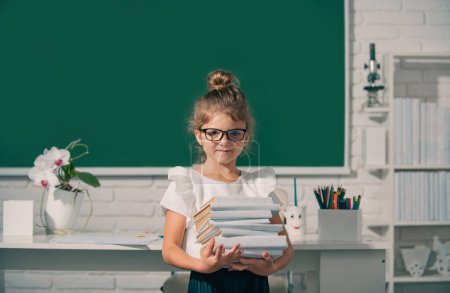 Foto de Cute child at school. Kid is learning in class on background of blackboard. Nerd school girl in glasses with books on blackboard - Imagen libre de derechos