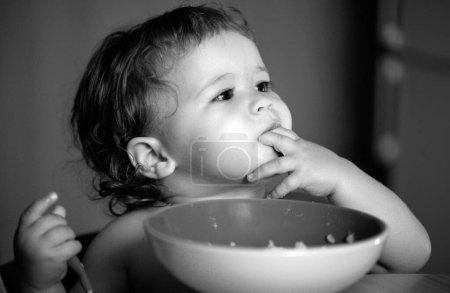 Photo for Funny little baby in the kitchen eating with fingers from plate. Child nutrition concept - Royalty Free Image
