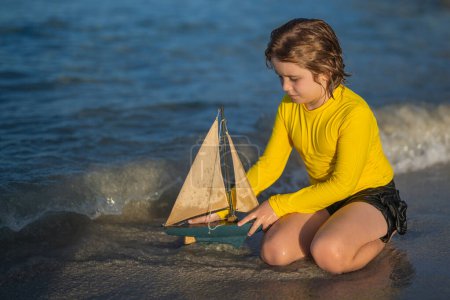 Téléchargez les photos : Jeu d'enfant avec bateau jouet dans les vagues de la mer à la plage pendant les vacances d'été. Vacances d'enfance et d'été en famille. Rêve en voyage. Visite guidée en voilier. Concept de rêve pour enfants. Bon été - en image libre de droit