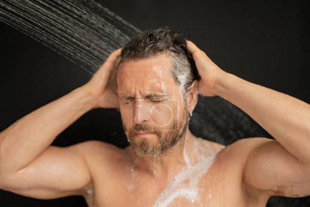 Téléchargez les photos : Homme hispanique laver les cheveux dans la salle de bain. Pommeau de douche dans la baignoire. Visage masculin sous la douche. Un homme prend une douche dans la salle de bain. Un mec qui se douche. Concept de salle de bain. L'homme est sous l'eau gouttes dans la salle de bain - en image libre de droit