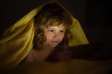 Téléchargez les photos : Enfant au lit sous une couverture joue sur une tablette dans un jeu dans le noir. Le visage des enfants est éclairé par une tablette. Enfant utilisant une tablette au lit à la maison. Enfant dans la chambre à coucher regardant un film ou jouant au jeu - en image libre de droit